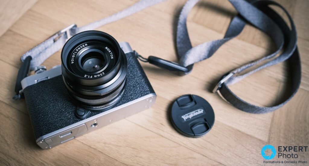 Voigtländer Nokton 23mm f/1.2 sur boîtier Fujifilm X, vue en plongée sur parquet, bouchon Voigtländer et sangle textile posés à côté