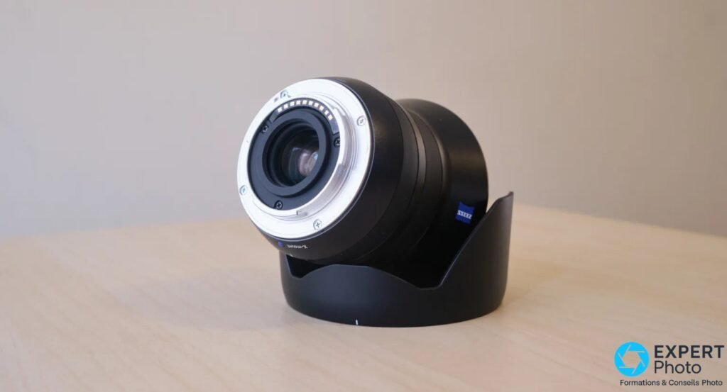 Zeiss Touit 2.8/12, vue 3/4 arrière, monture X-mount argentée avec contacts électriques visibles, logo Zeiss bleu sur le fût