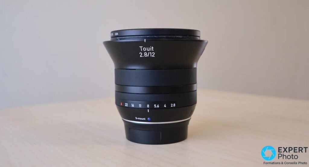 Zeiss Touit 2.8/12, vue frontale, bague de diaphragme A-22-16-11-8-5,6-4-2,8, X-mount visible, pare-soleil, fond bois