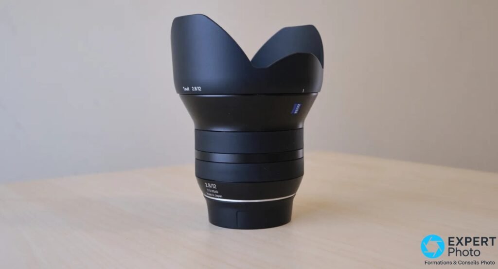 Zeiss Touit 2.8/12 debout avec pare-soleil tulipe, gravure 2.8/12, logo Zeiss bleu sur le côté, fond bois clair